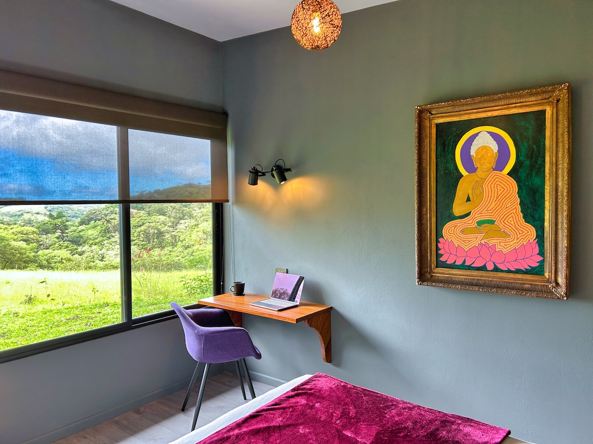 A serene bedroom is featured with a calming colour palette. A wall-mounted artwork of a seated figure is positioned above a small desk with a chair. A large window offers a view of the lush greenery outside, allowing natural light to illuminate the space.