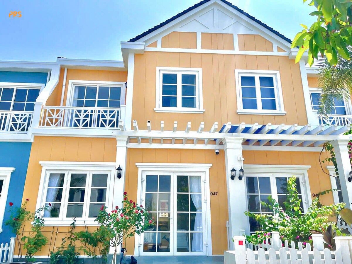 The exterior of a two-story house is showcased, featuring a warm yellow facade accentuated by white trim. A covered porch with decorative railings invites relaxation. Greenery and flowers surround the entrance, adding a touch of nature to the welcoming atmosphere.
