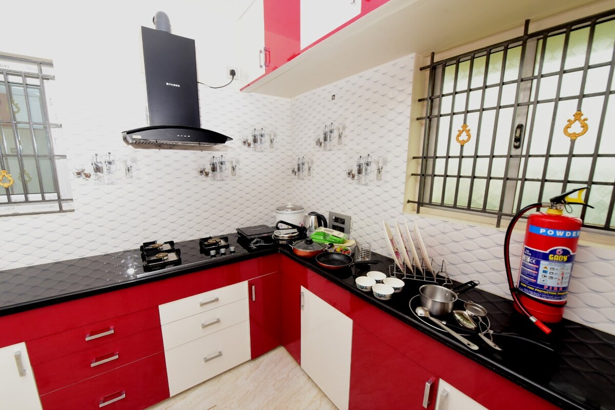 The kitchen features a mix of red and white cabinetry with a sleek black countertop. Two gas burners are installed, accompanied by various cooking utensils and containers. A modern exhaust hood is mounted above, and a fire extinguisher is visible for safety.
