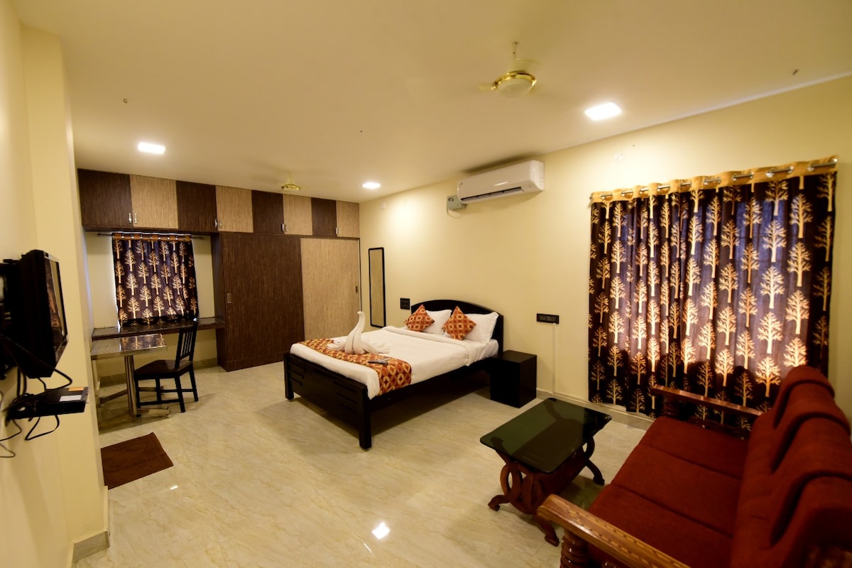 A spacious room features a large bed adorned with decorative pillows. A seating area with a red sofa is positioned beside a dark wooden table. Natural light filters through patterned curtains, illuminating the room's neutral tones and modern furnishings. A flat-screen TV is mounted on the wall.