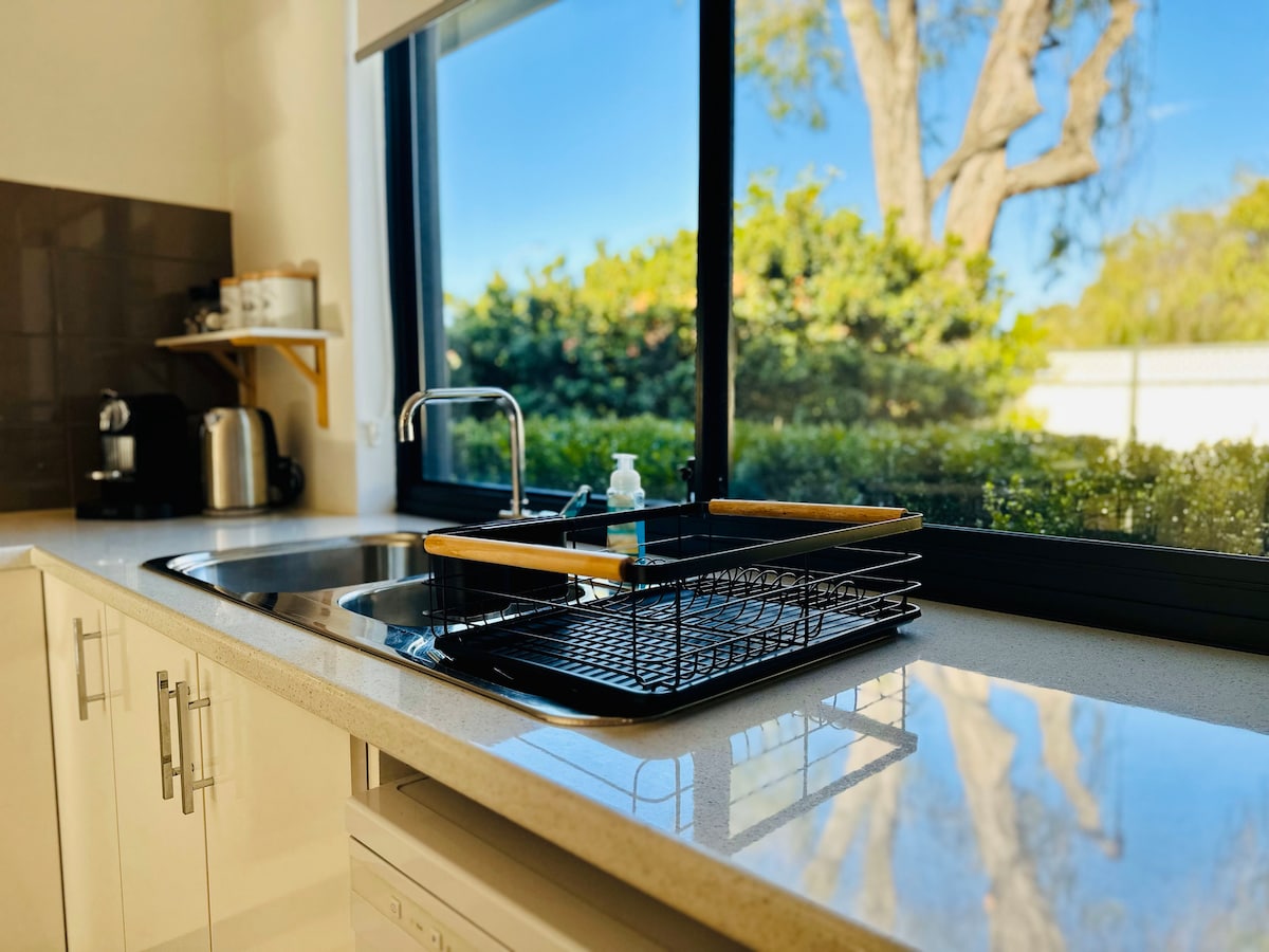 A modern kitchen space features sleek white cabinetry and a large window offering views of greenery. A neatly arranged dish drying rack sits on the countertop beside a stainless steel sink, with a kettle and various kitchen supplies visible in the background.