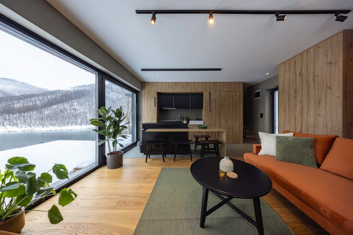 A contemporary living space features large windows framing serene lake views. A wooden dining table and kitchen area are visible in the background. A cozy orange sofa complements a dark round coffee table, with greenery and a decorative item placed on the table, enhancing the inviting atmosphere.