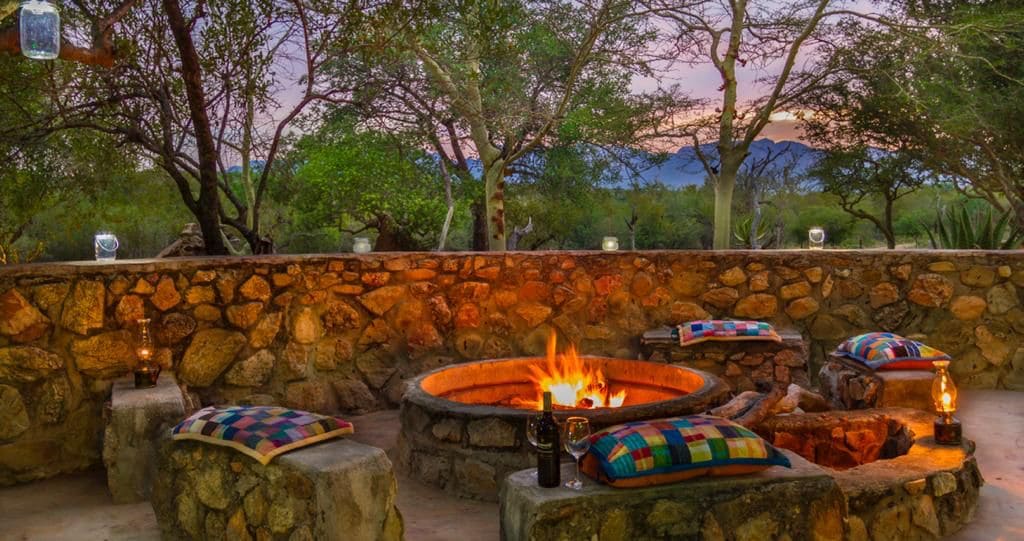 A circular stone fire pit sits surrounded by rustic seating, featuring colorful cushions. Lanterns provide soft illumination as the sun sets behind distant mountains, highlighting the lush greenery of the surrounding area.
