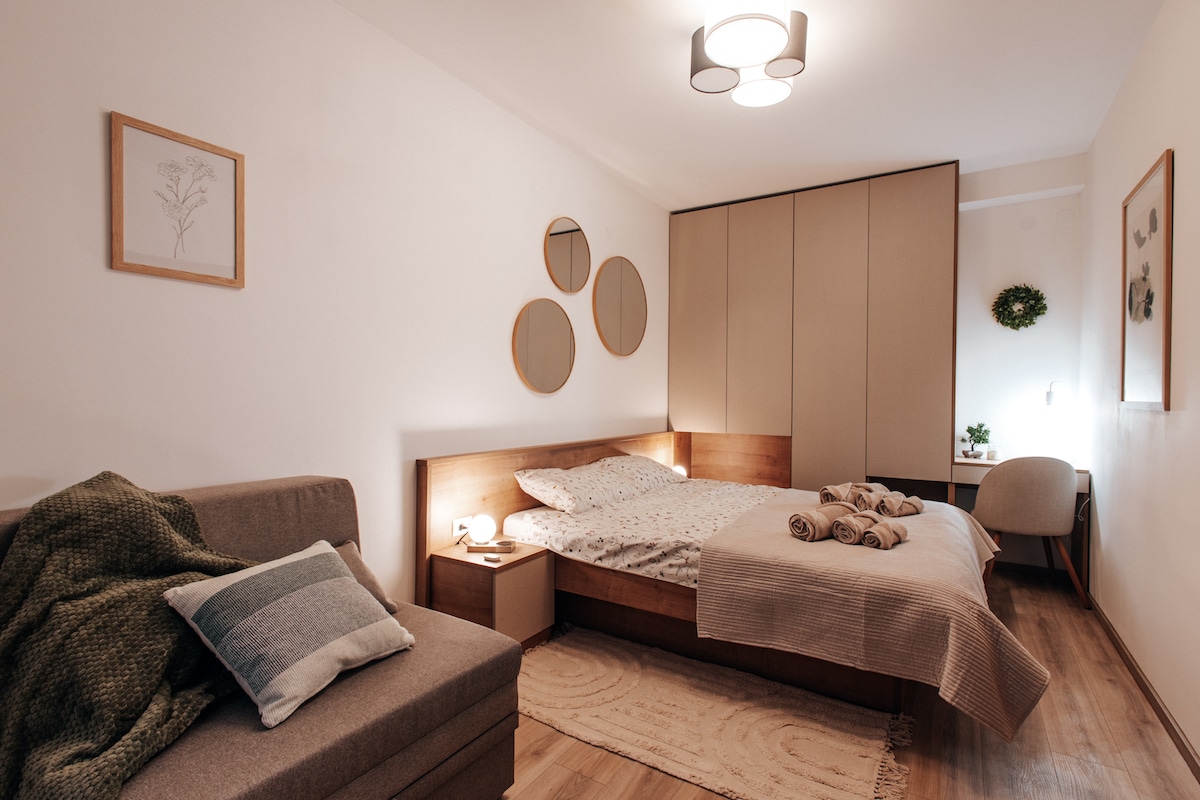 The bedroom features a spacious bed with neutral-toned bedding, complemented by decorative pillows. A large wardrobe provides ample storage. Circular mirrors adorn the wall, reflecting soft light, while a cozy armchair and a small sofa are positioned nearby, enhancing the room's comfort.