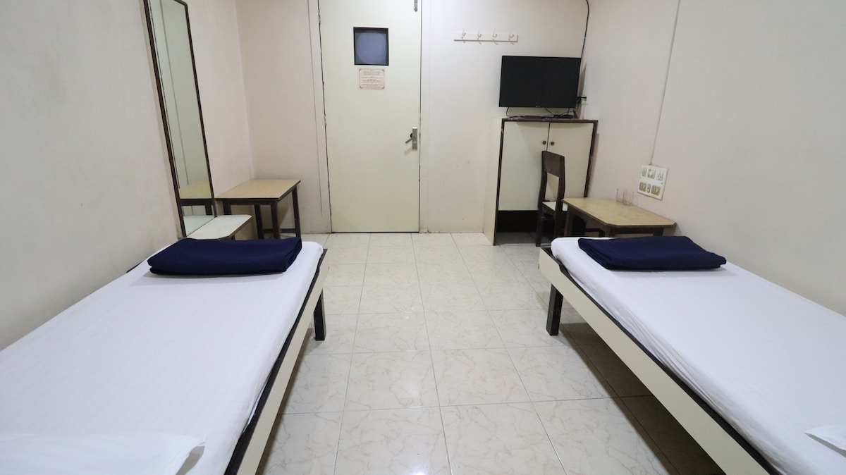 The room features two single beds with dark blue beddings, positioned parallel to each other. A small wooden table and chair are present against one wall, while a flat-screen TV is mounted nearby. A mirror adds to the spacious appearance, and a door leads to the hallway.