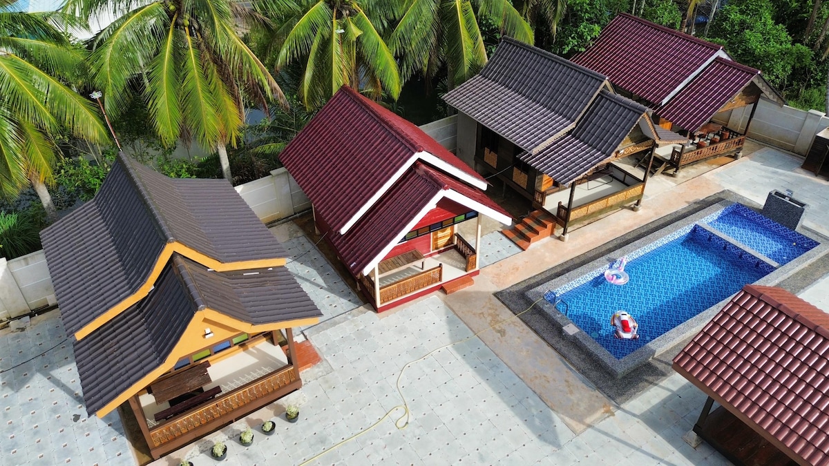 A bird's-eye view captures multiple homestay units featuring sloped roofs in warm hues. The central swimming pool, surrounded by palm trees, is adorned with colorful floats. A spacious patio area offers ample room for relaxation and gathering.