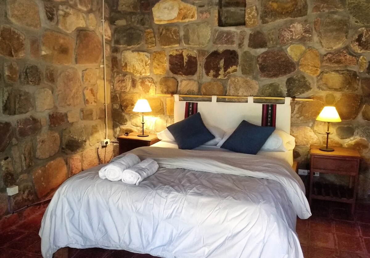 A cozy bedroom features a stone wall backdrop with softly lit bedside lamps on wooden nightstands. A spacious bed with a light blanket and two blue pillows is centrally positioned. Rolled towels are neatly arranged on the bed for guest convenience.