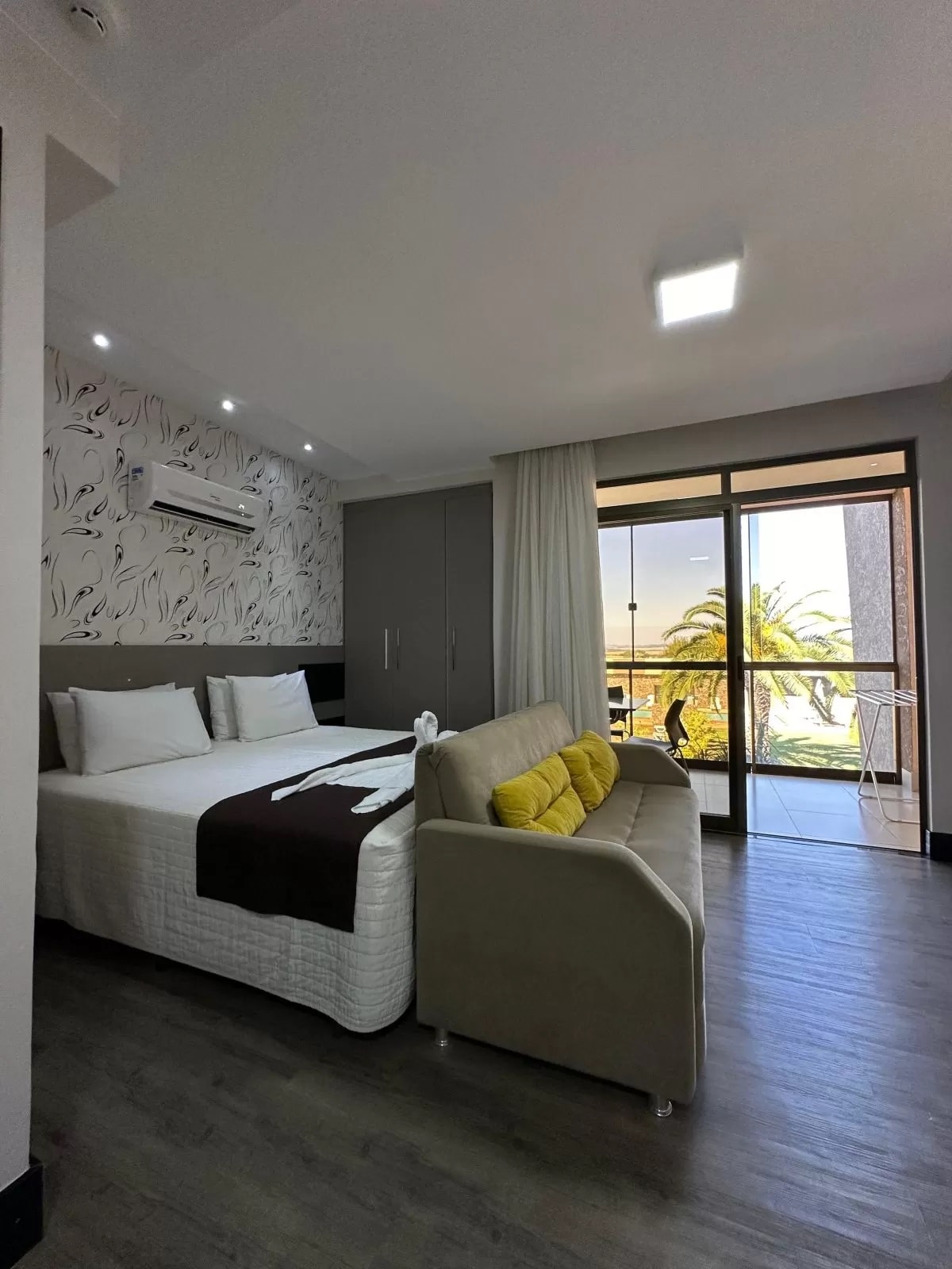 A spacious room features a large bed with white linens and decorative pillows. A cozy sofa in neutral tones is positioned near a sliding glass door, allowing natural light to illuminate the space. A balcony with views of palm trees is accessible from the room.