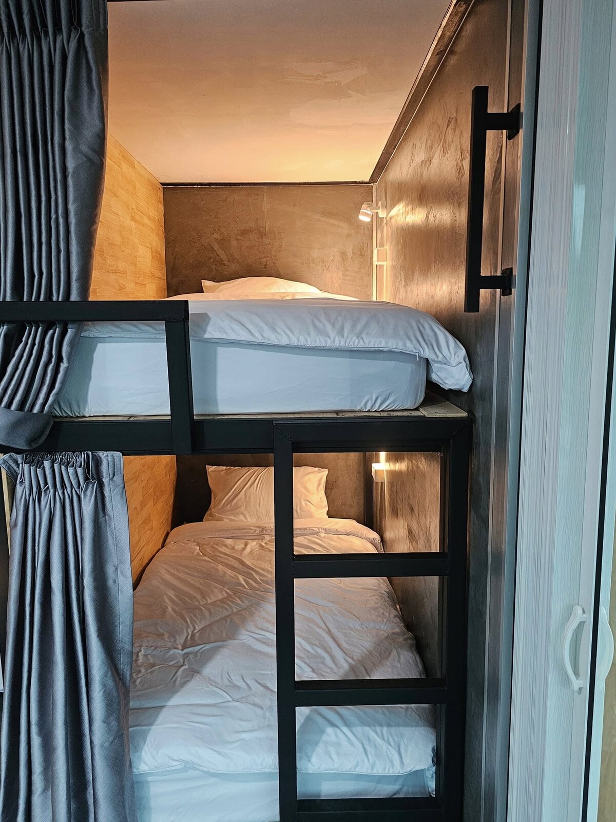 A compact sleeping area features a double-decker bed with a spacious upper bunk and a lower sleeping space. Light fixtures on both levels provide illumination, while gray curtains offer privacy. The walls are designed with a minimalist aesthetic, enhancing the room's cozy feel.