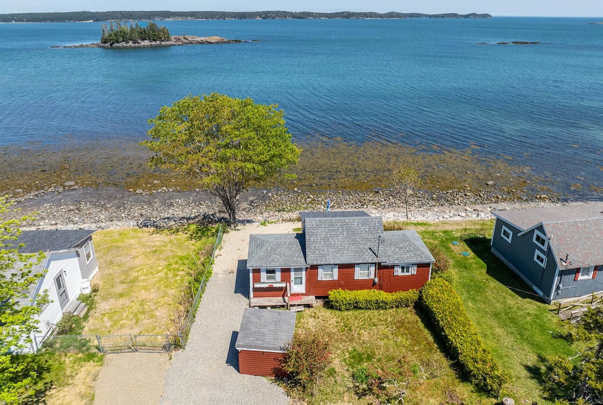 A cottage is positioned near the shoreline, with a clear view of the water and rocky beach. Surrounding greenery provides a natural contrast to the red facade. An island is visible in the distance, and neighboring cottages are seen along the coast.