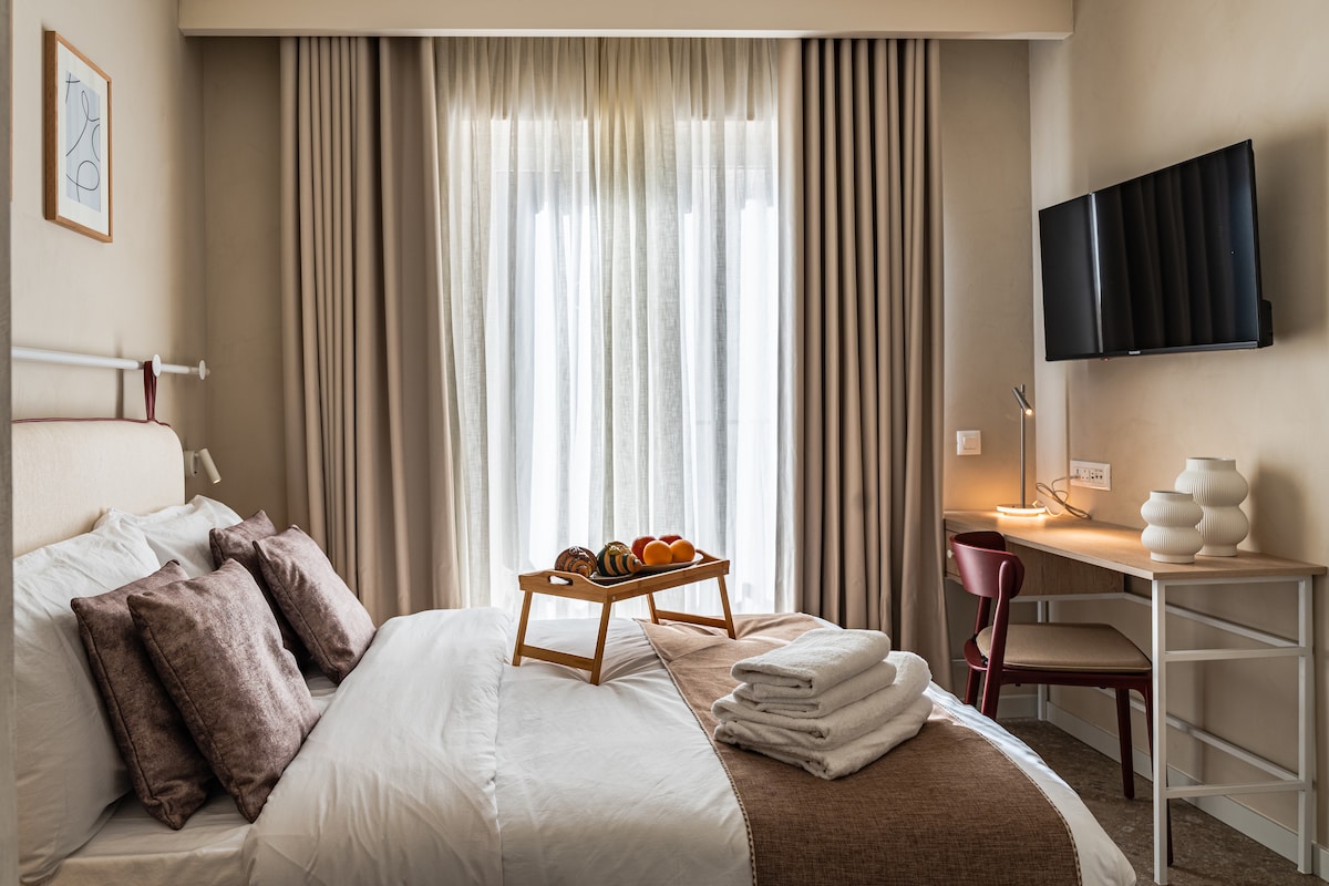 A well-appointed sleeping area features a spacious bed dressed with light linens and an assortment of decorative pillows. A tray with fruits rests on the bed, while a desk with a lamp and a flat-screen TV are positioned nearby. Sheer curtains allow soft light to filter in.