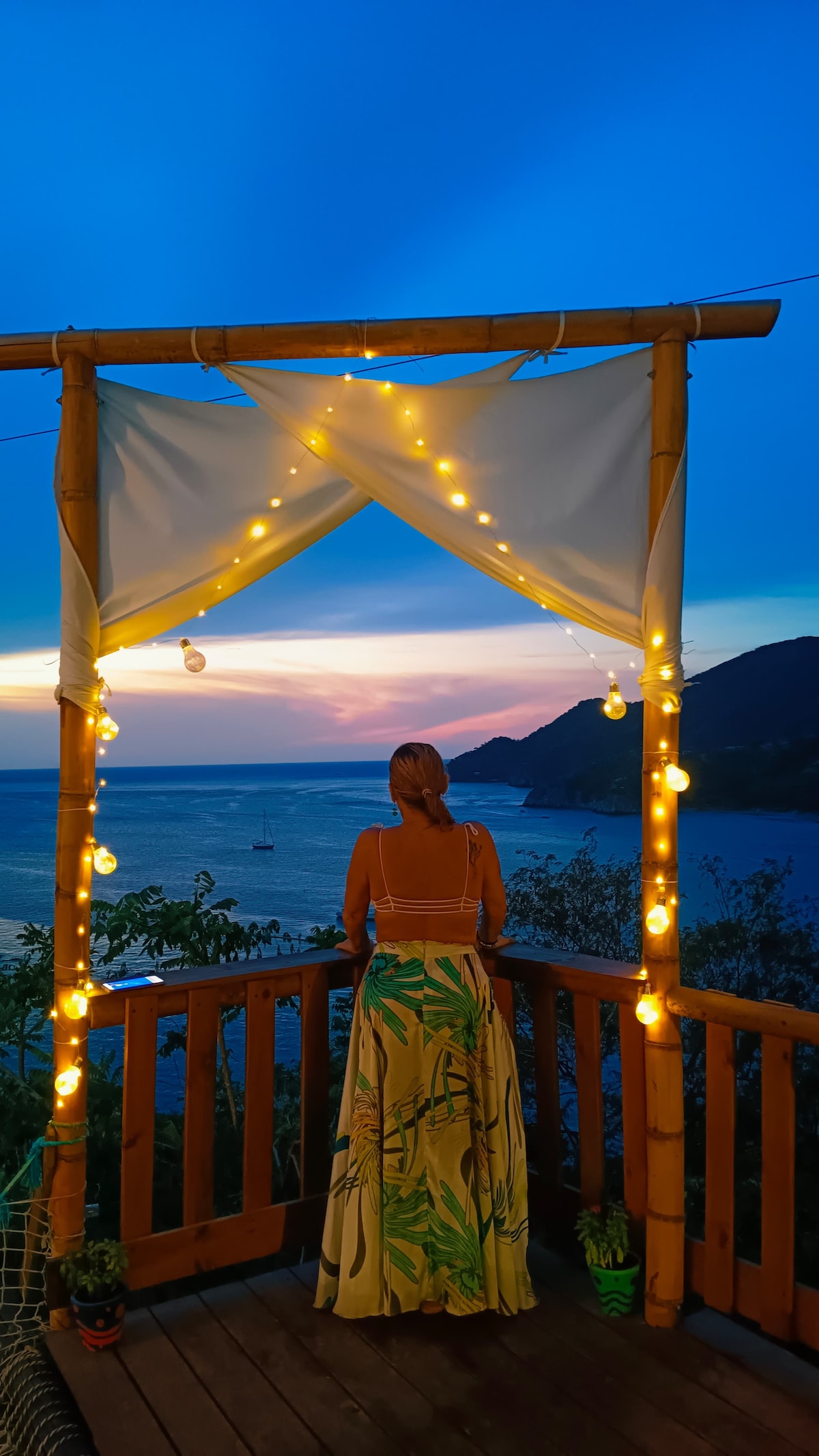 A wooden terrace adorned with soft lighting features a draped canopy. The view showcases a serene ocean landscape under a vibrant sky at sunset. Lush green foliage contributes to the tranquil setting, inviting relaxation and contemplation.