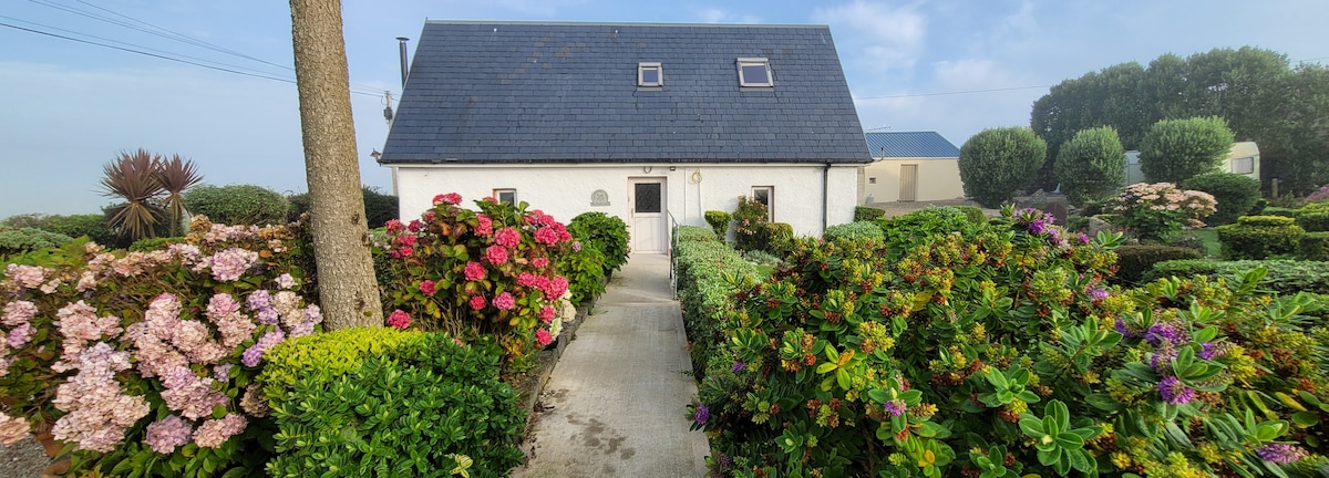 A charming cottage is framed by vibrant flowering bushes and lush greenery. A concrete path leads to the front door, surrounded by colorful blooms in shades of pink and green. The cottage features a slate roof and large windows, creating a welcoming entrance.