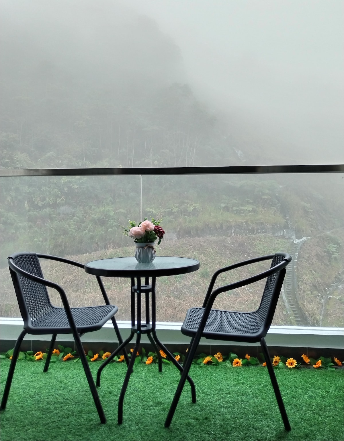 An outdoor seating area features a small round table with a vase of flowers, accompanied by two black chairs. A lush green carpet creates a cozy feel, while fog-covered mountains and greenery can be seen in the background.