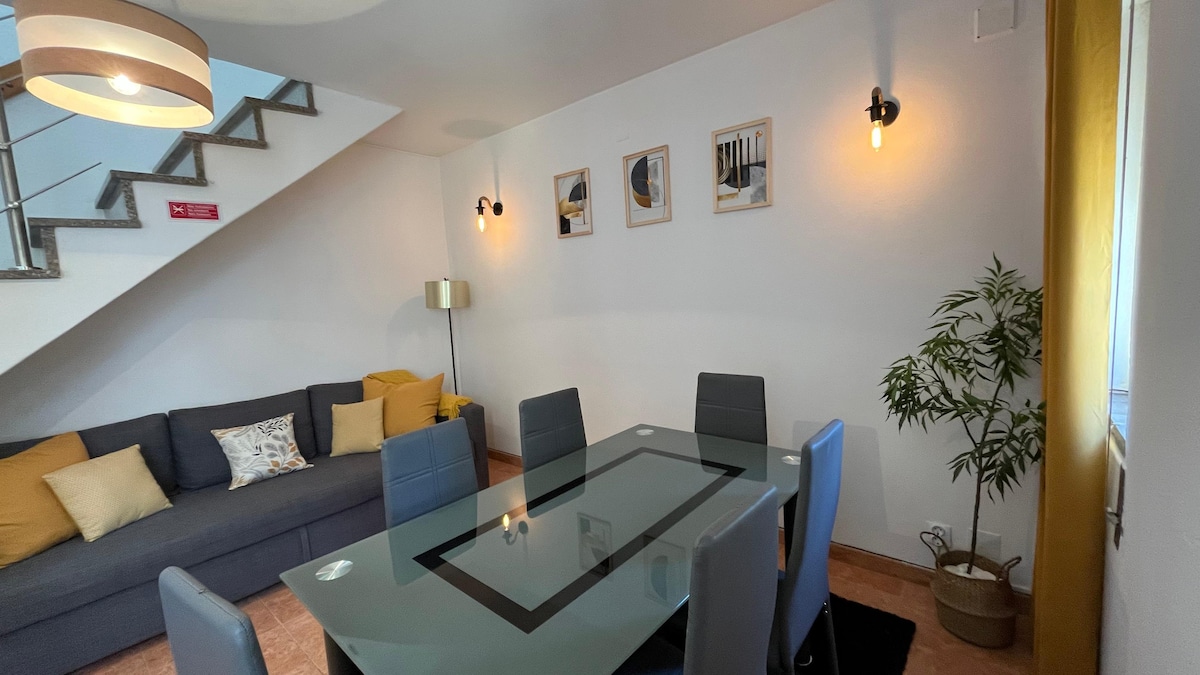 A bright dining area features a glass table surrounded by six gray chairs. A cozy sofa with yellow and patterned cushions is positioned against the wall. Warm lighting is provided by wall-mounted fixtures and a pendant lamp, creating a welcoming space.