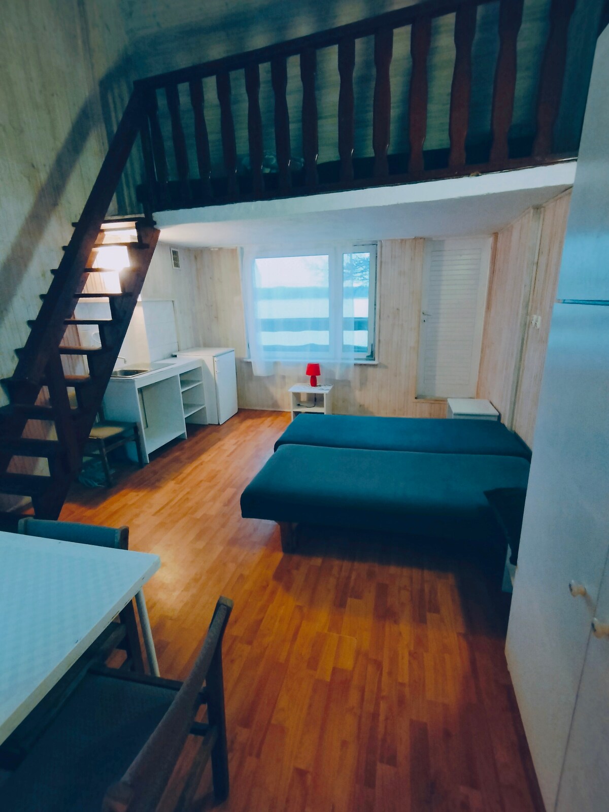 A cozy living area features a wooden staircase leading to a lofted space. Two twin beds are positioned near a window, allowing natural light to fill the room. A small dining table is visible, accompanied by chairs, against a backdrop of light-colored walls and warm wooden flooring.