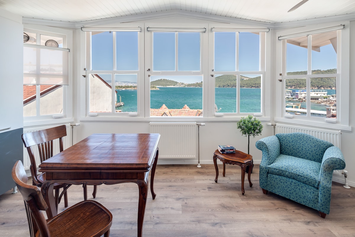 A bright living area features large windows allowing scenic views of the sea and the surrounding landscape. A wooden dining table with matching chairs is positioned beside a blue upholstered couch. Natural light fills the space, enhancing the inviting atmosphere.