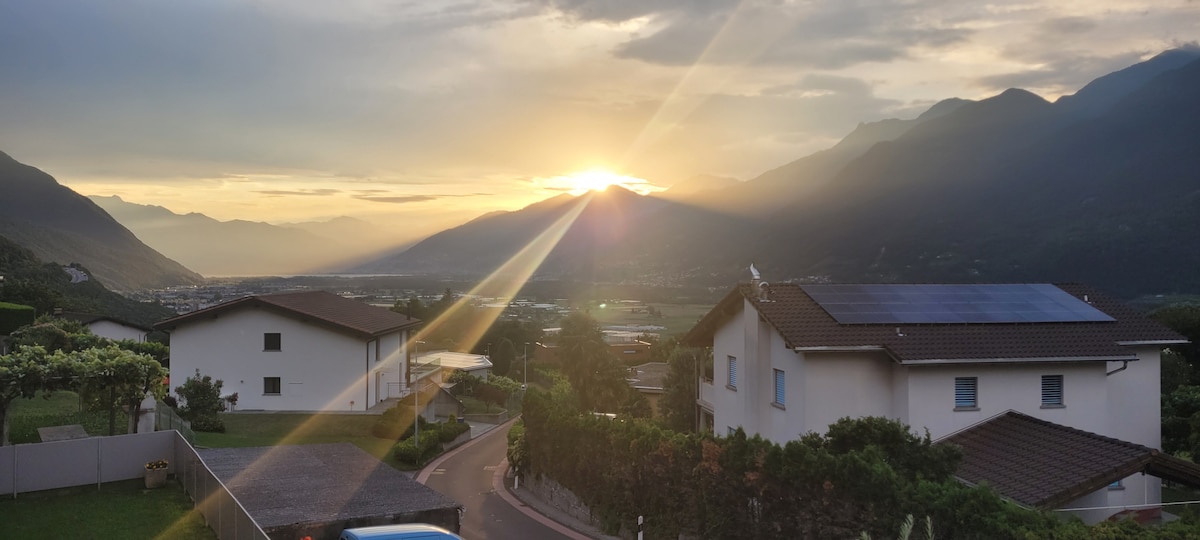 A serene landscape features the sun setting behind mountains, casting warm rays over the valley. Two nearby houses with light-colored façades are visible, along with green hills and gently winding roads, providing a tranquil view of the surrounding natural beauty.