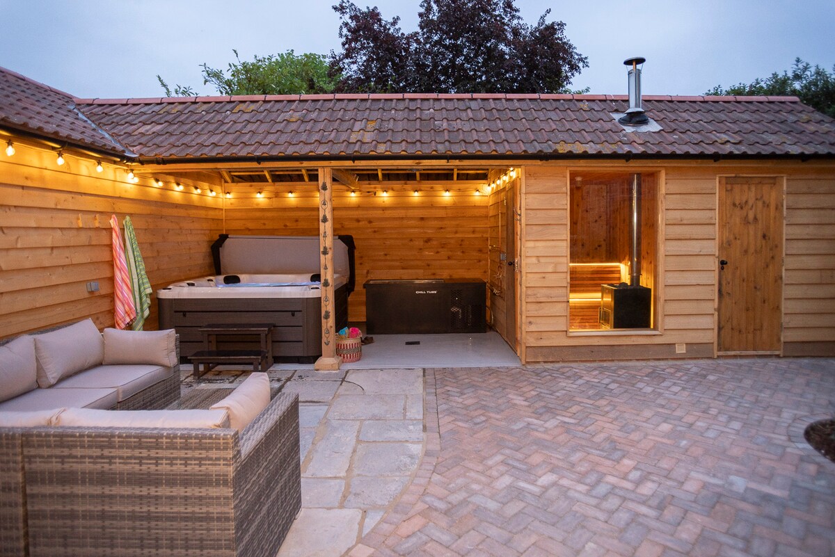 A private courtyard wellness suite features a hot tub and a sauna, with an outdoor shower nearby. Soft lighting creates an inviting ambiance. Comfortable seating is arranged around a stone-paved area, and wooden walls add warmth to the space.