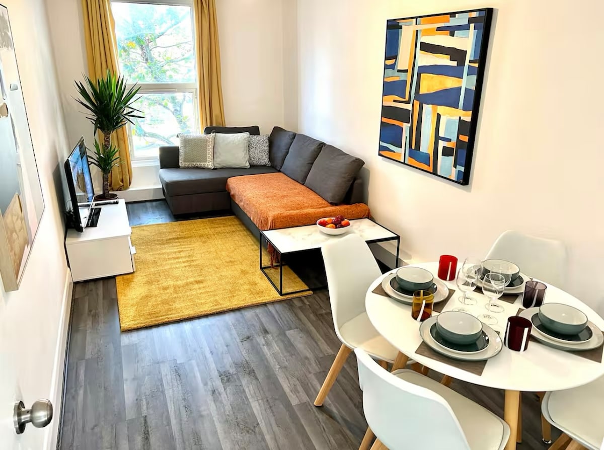 A bright living area features a gray sofa with an orange chaise, complemented by a textured area rug. A small dining table is set for four, with dinnerware neatly arranged. A vibrant piece of wall art adds a splash of color, and a large window allows natural light to enter.