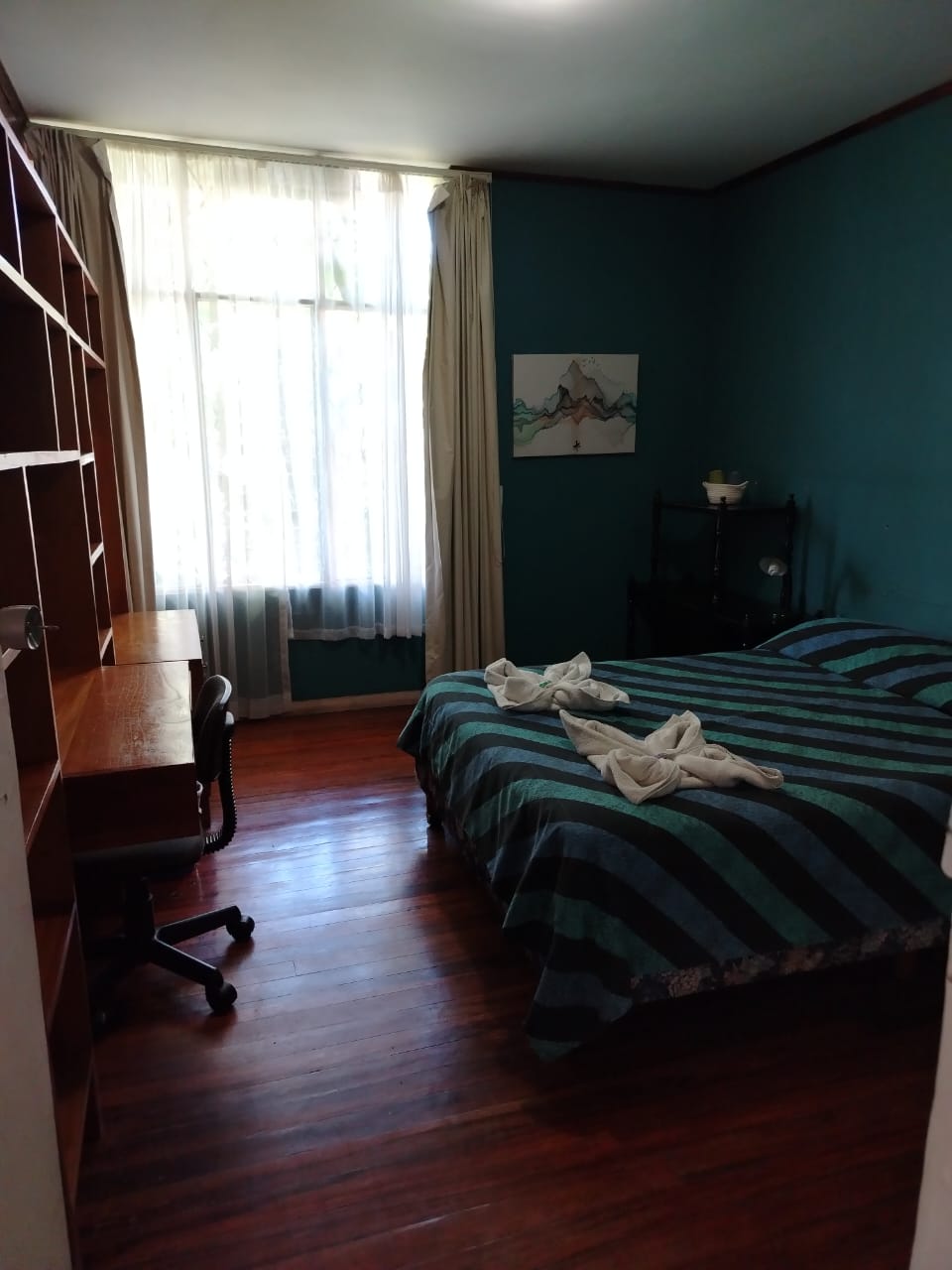 A welcoming sleeping area features a spacious bed with striped bedding. A desk is positioned alongside the window, allowing natural light to brighten the room. Light curtains gently filter sunlight, and wooden flooring adds warmth to the space.