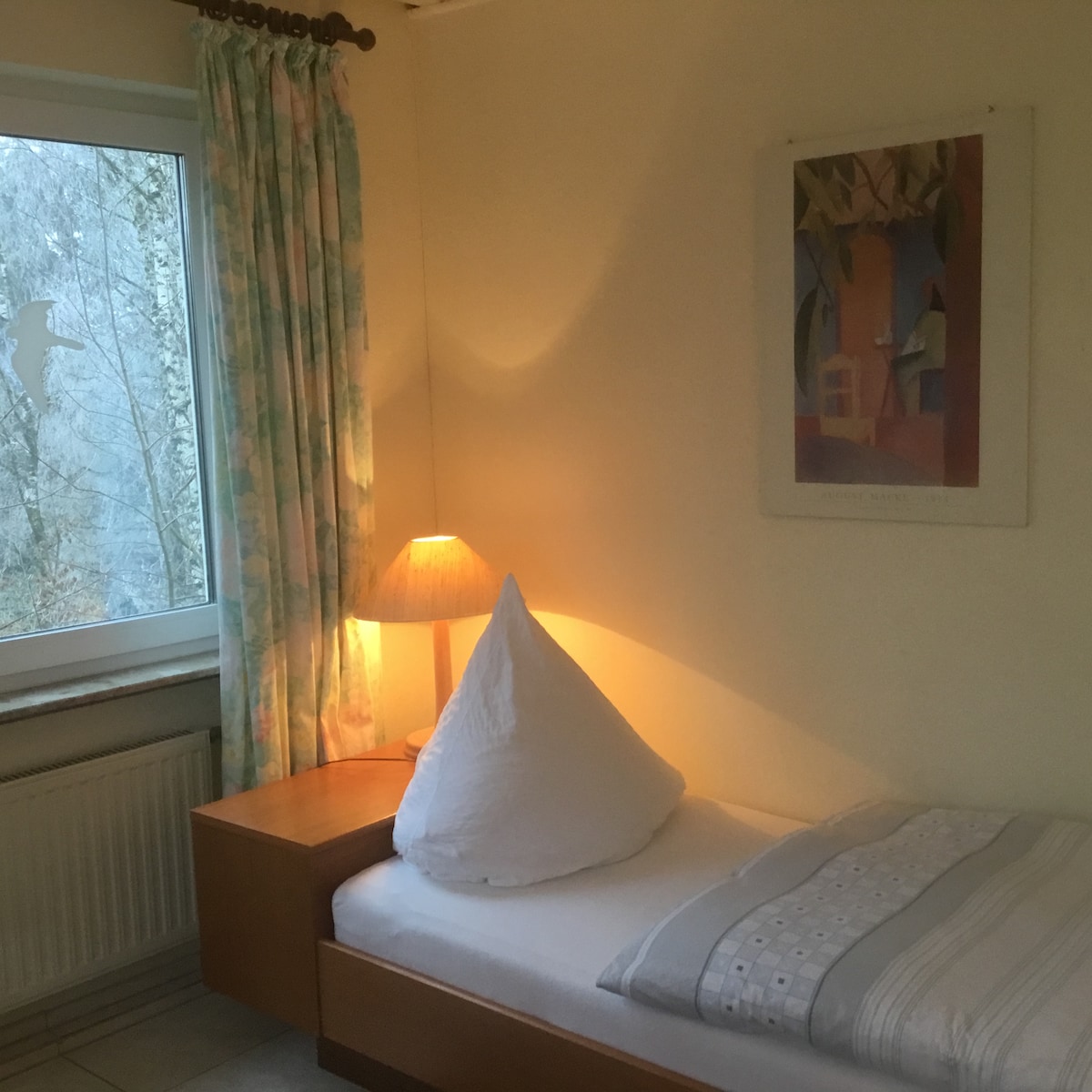 A cozy sleeping area features a single bed topped with a triangular pillow and a soft blanket. A bedside table holds a warm lamp, casting a gentle glow. Light-colored curtains frame the window, allowing natural light to brighten the room, while a piece of abstract wall art adds a subtle touch.