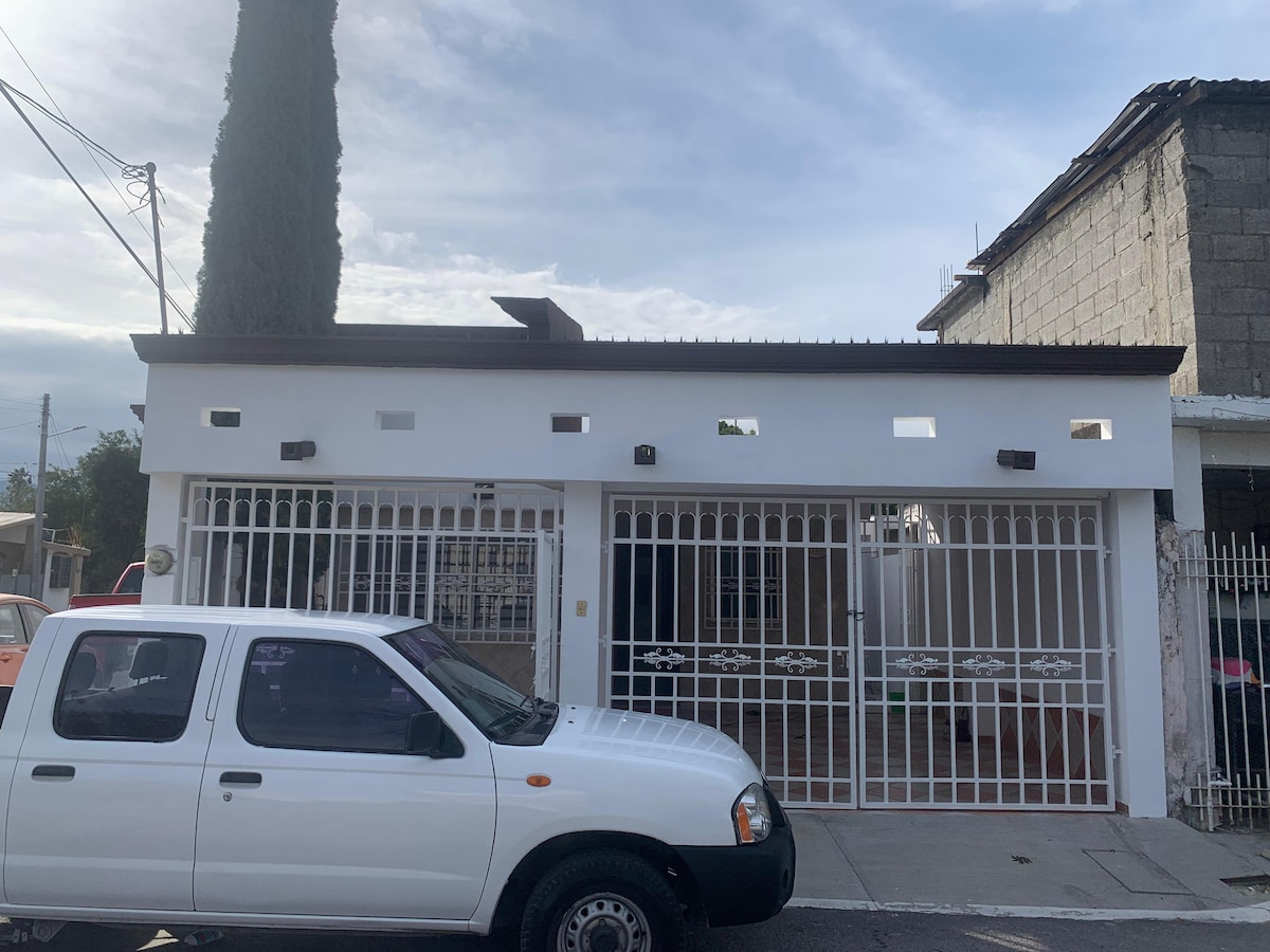 A modern white facade with a simple design is displayed, featuring a gated entrance and several small windows along the top. A white pickup truck is parked in front of the building, contributing to the casual atmosphere of the neighborhood.