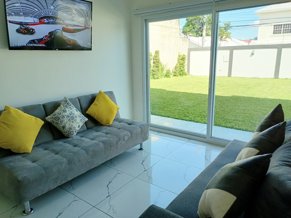 A bright living area features a large glass door that opens to a grassy outdoor space. Two gray sofas with colorful cushions are arranged invitingly. Natural light fills the room, highlighting the sleek tile flooring and a mounted television.