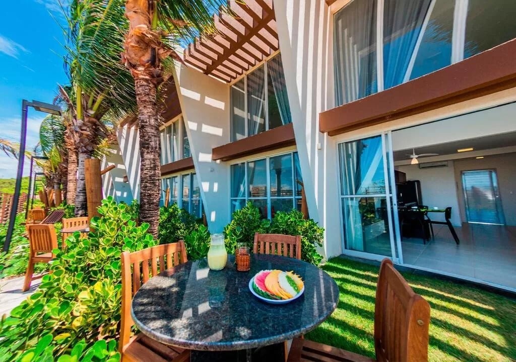 An outdoor seating area is presented with a round stone table and wooden chairs surrounded by lush greenery. Bright sunlight filters through the leaves of nearby palm trees, creating a relaxing ambiance. A plate of colorful food is placed on the table, inviting enjoyment on the patio.
