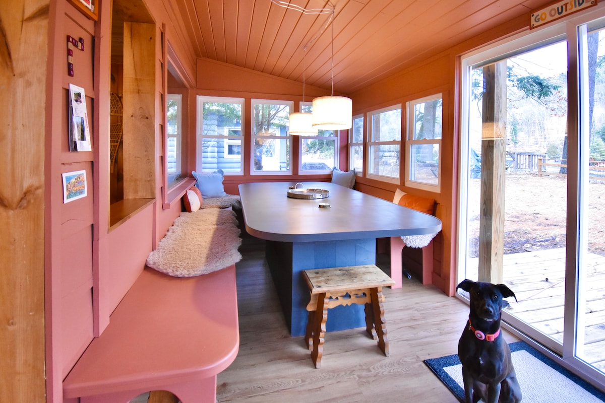 A bright dining area features large windows allowing natural light to flood the space. A long wooden table is surrounded by cozy seating. A decorative light fixture hangs above, and a dog stands on a welcome mat near an outdoor entrance.