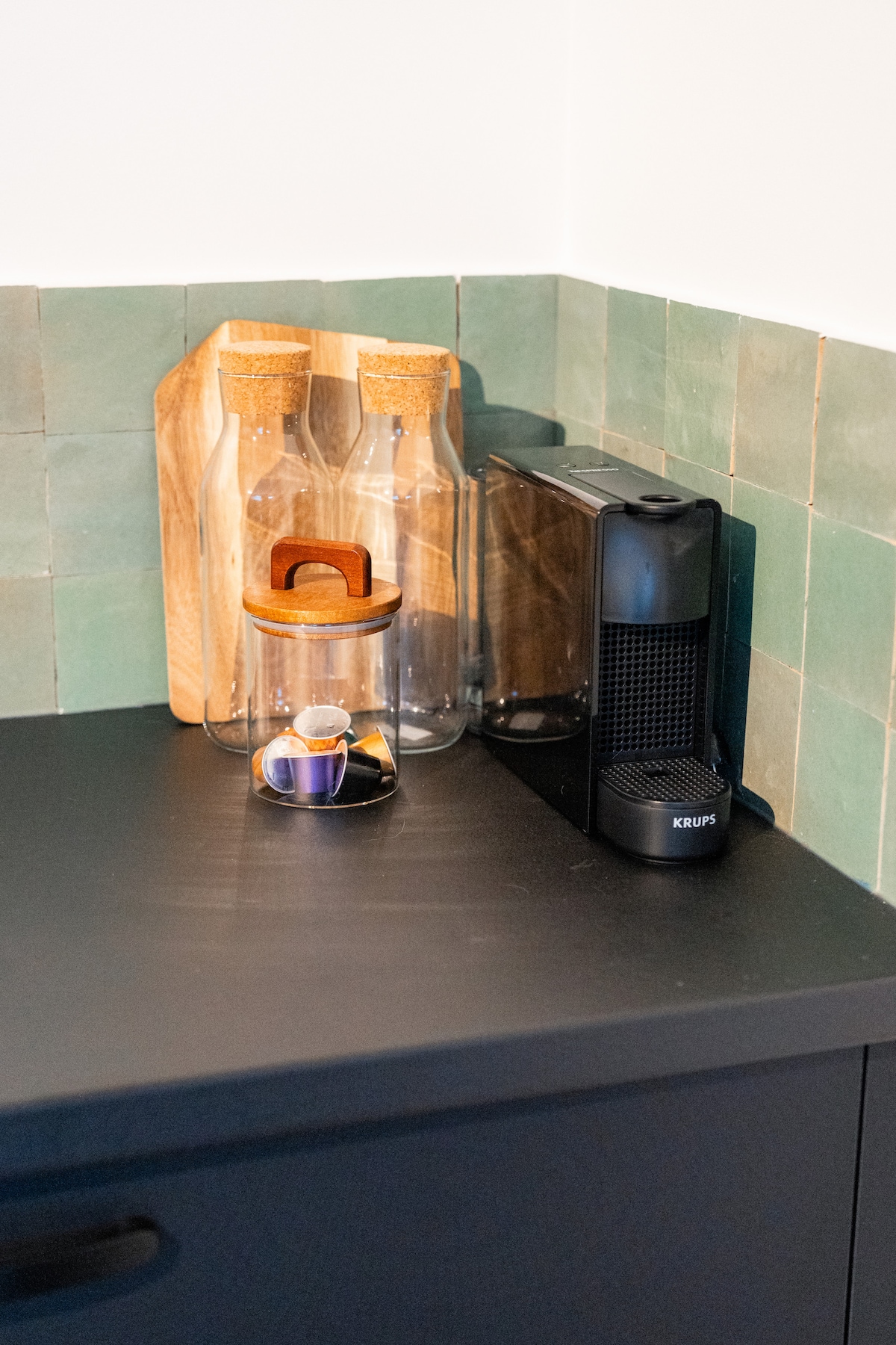 A neatly arranged kitchen corner features two glass bottles with cork stoppers, positioned next to a wooden cutting board. A sleek coffee machine sits on the dark countertop, accompanied by a container for coffee capsules, providing a blend of functionality and style.
