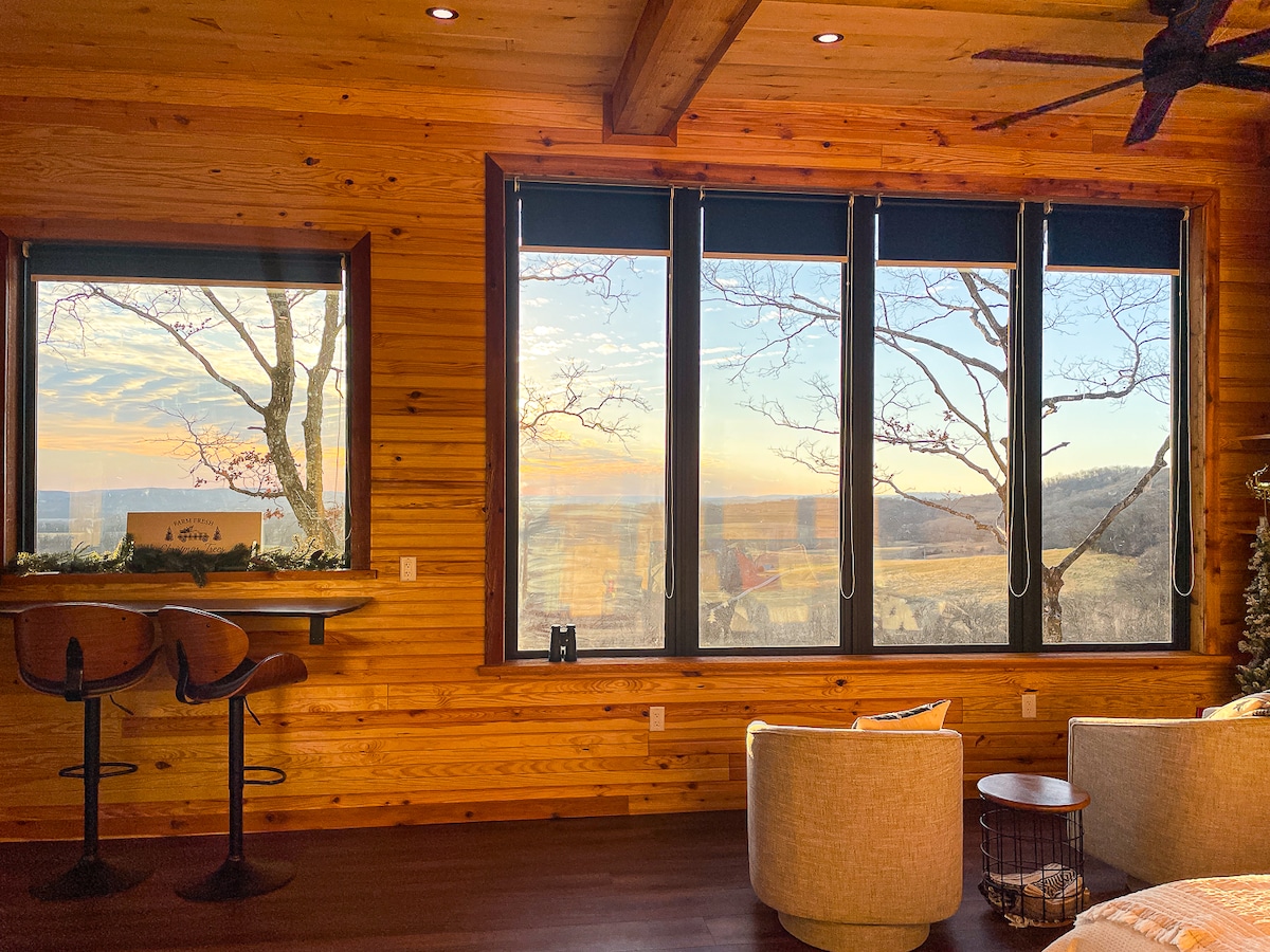 A cozy interior features large windows showcasing expansive views of the surrounding landscape. Wooden walls add warmth to the space, while two bar stools and a stylish chair provide seating options. Light streams in, reflecting off surfaces and enhancing the natural ambiance.