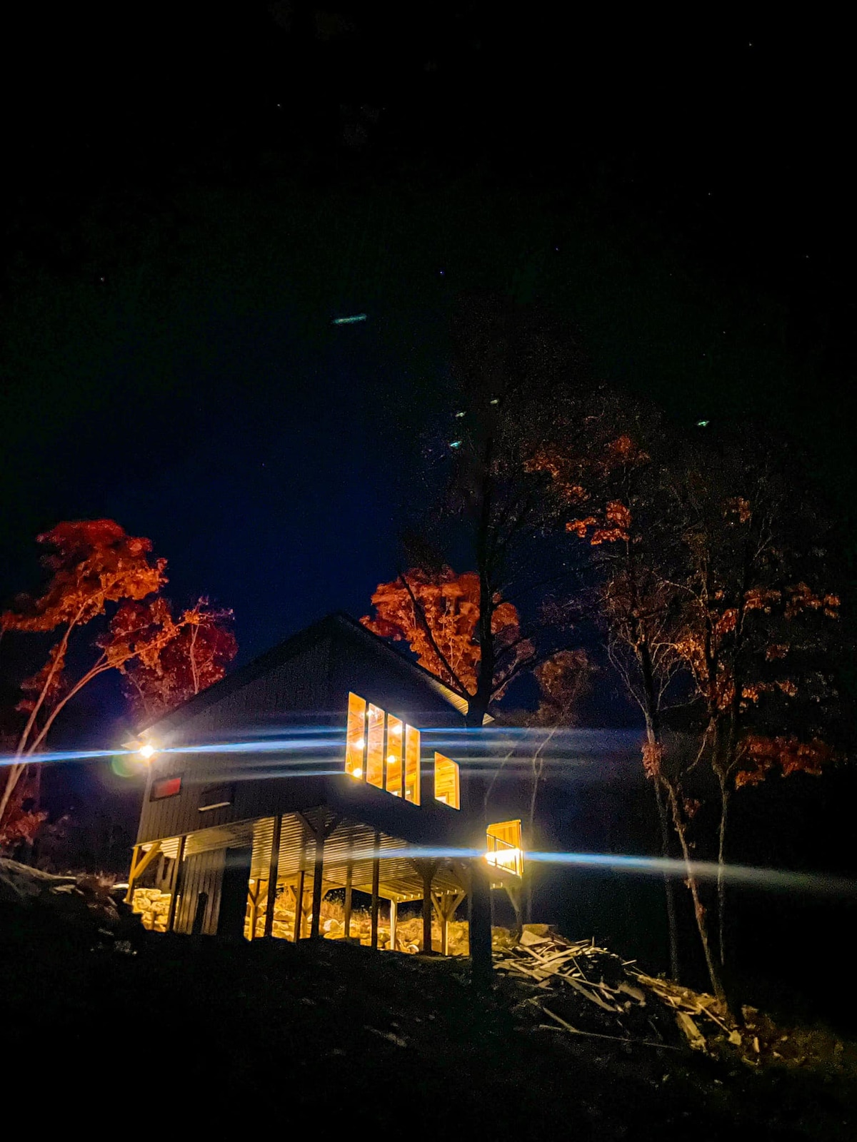 The treehouse is illuminated at night, highlighting its modern architecture nestled among trees. Warm lights glow from the windows, contrasting with the dark sky. Surrounding foliage displays vibrant orange hues, adding a serene ambiance to the outdoor setting.