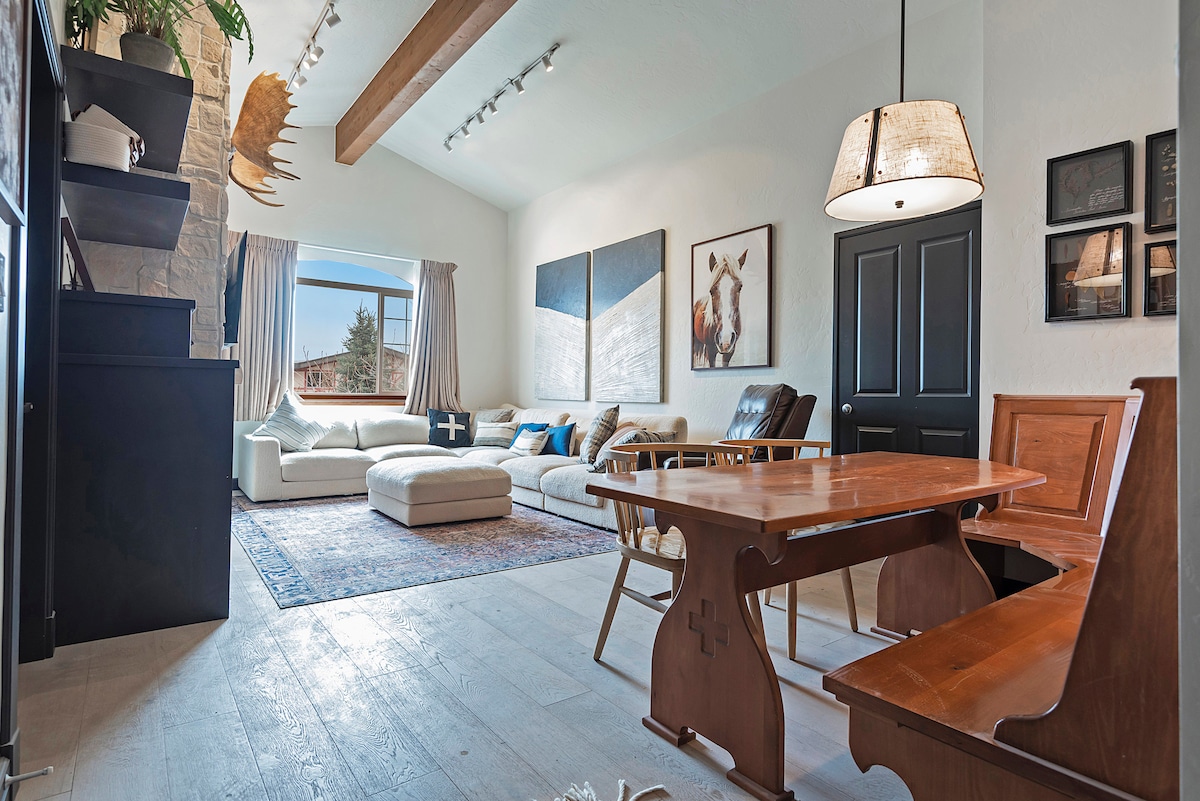 A spacious living area is highlighted by a large sectional sofa, accompanied by a decorative rug. Natural light enters through a window, illuminating artwork on the walls. A dining nook with a wooden table and bench seating offers a cozy space for meals.