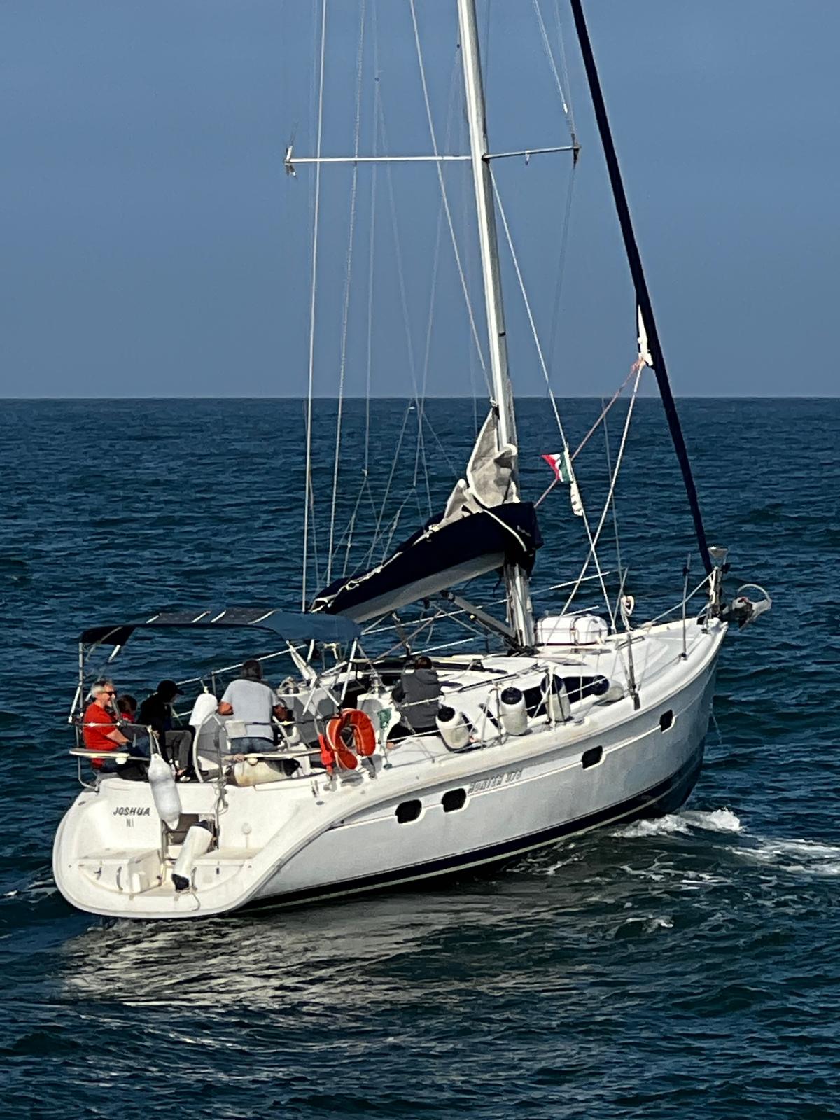 A white sailing yacht is seen navigating the open water, with several individuals on board. The sail is raised, and a canopy provides shade over the cockpit area. The calm sea creates a contrasting backdrop against the vessel's sleek design.
