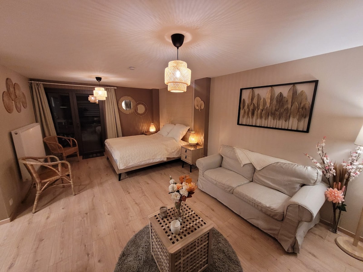 A well-furnished studio features a spacious bed adorned with neutral bedding, paired with a light-colored sofa and a decorative coffee table. Soft lighting is provided by pendant lamps, and large glass doors lead to a private terrace, creating a cozy atmosphere.