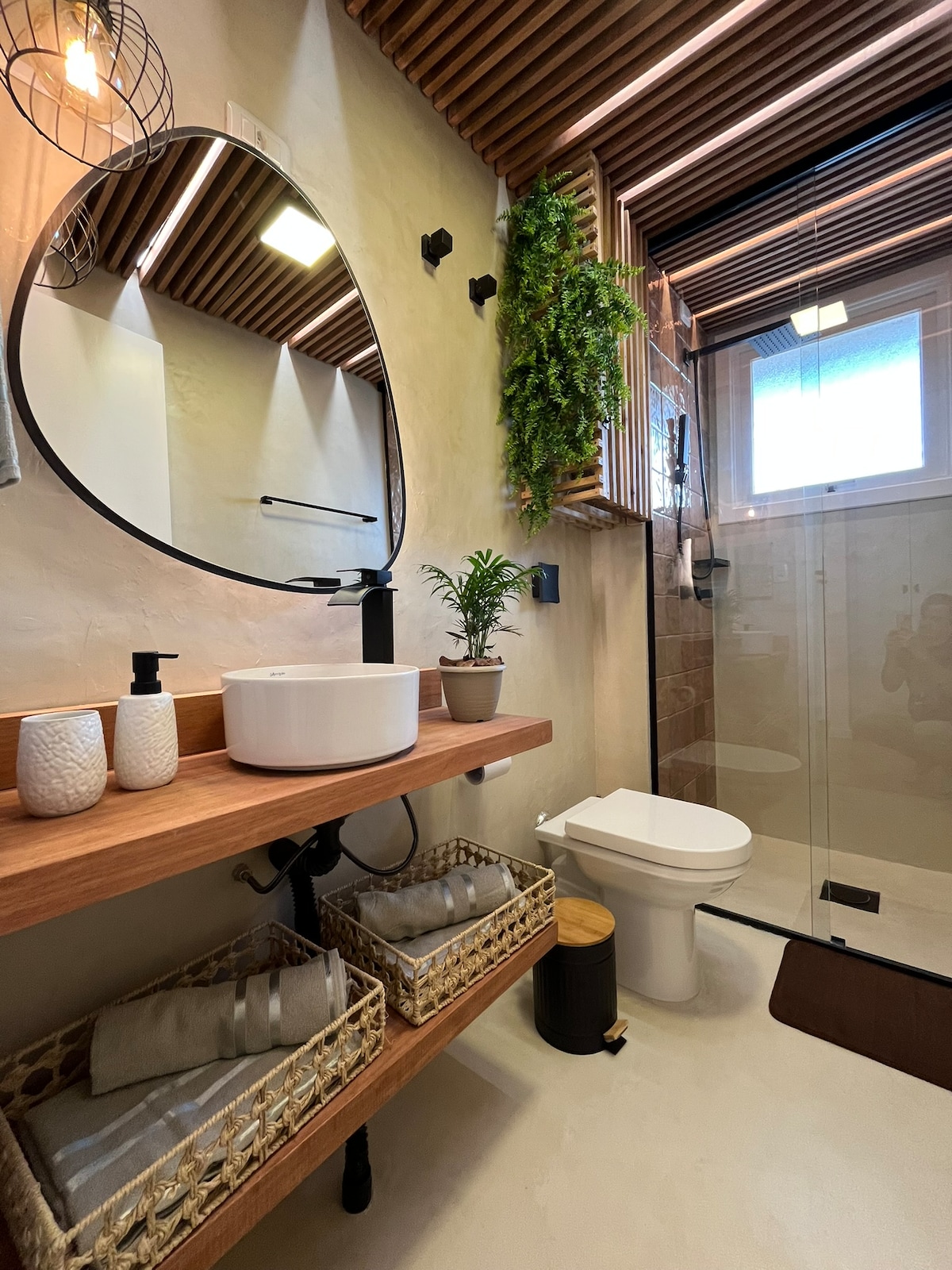 The bathroom features a modern design with a round mirror above a wooden shelf. A countertop basin is positioned alongside two woven baskets containing towels. A glass shower enclosure is visible in the background, with natural light filtering in through a window.