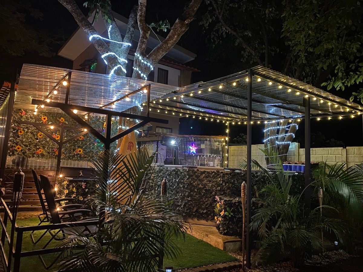 An outdoor relaxation area is displayed, featuring two covered seating arrangements under illuminated canopies. String lights create a soft glow, complemented by vibrant flowers and lush greenery. A glimpse of a pool can be seen in the background, enhancing the tranquil atmosphere.