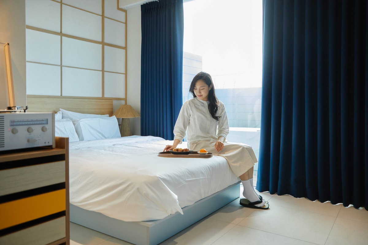 A spacious bed is neatly made with white linens, adorned with decorative pillows. A bedside lamp and a wooden nightstand with a retro-style music player are visible. A person is seated on the edge of the bed, holding a tray with refreshments, while blue curtains frame a large window.