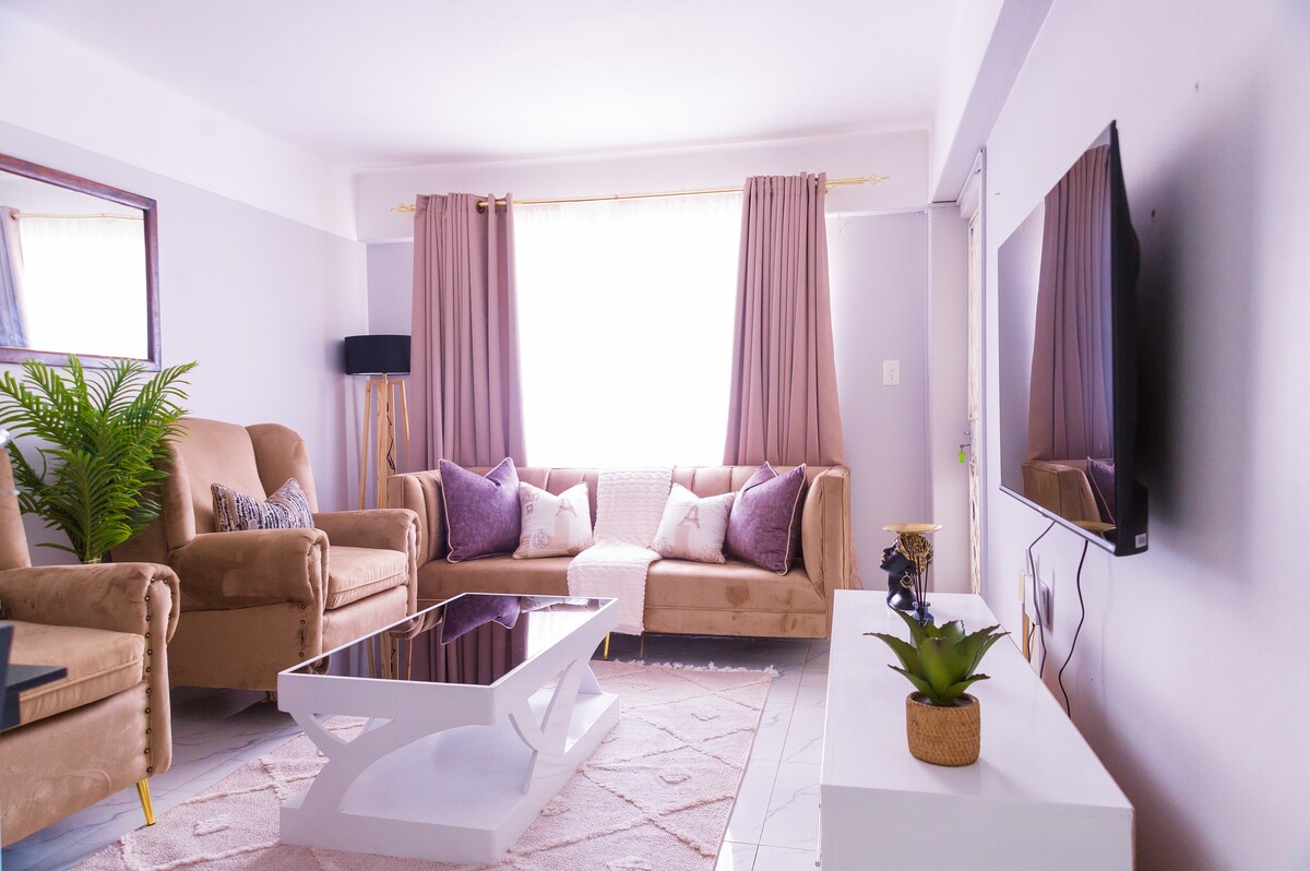 A modern living area features two plush armchairs and a comfortable sofa adorned with decorative pillows. A large coffee table rests on a soft area rug, while a flat-screen television is mounted on the wall opposite a window draped with elegant curtains.