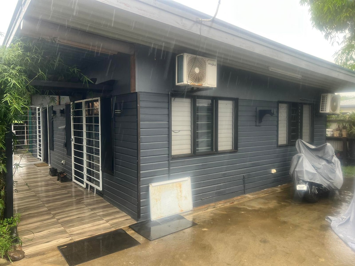 A modern single-story structure features a dark exterior with multiple windows. A covered wooden deck extends from the side, and air conditioning units are mounted on the walls. The space is surrounded by greenery, adding a natural element to the environment.
