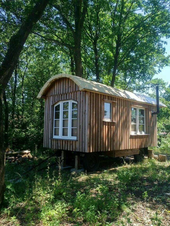 A cozy wooden caravan rests among lush greenery, featuring multiple windows that invite natural light. The curved roof adds a charming touch, while the surrounding trees provide a serene buffer from the outside world.