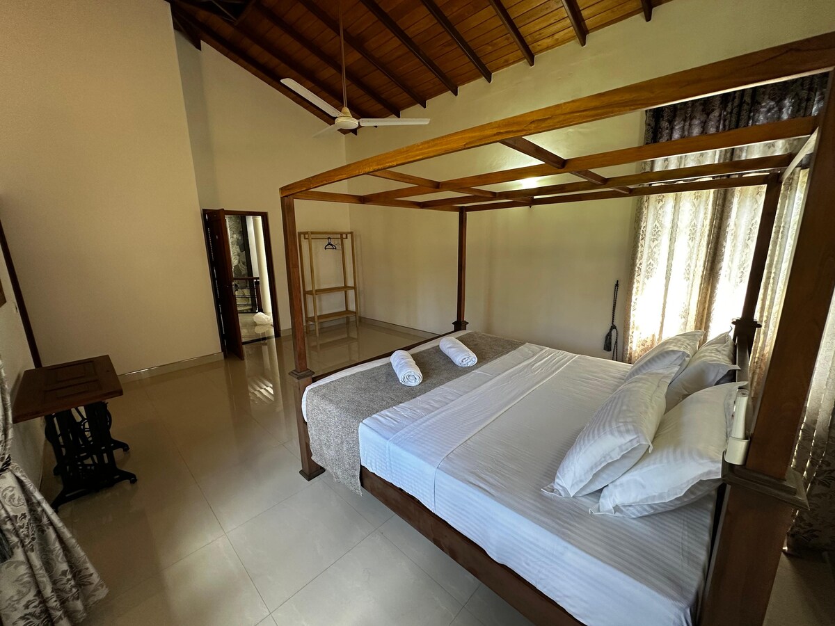 A spacious bedroom features a four-poster bed with a light-colored bedding arrangement. Natural light filters through sheer curtains, illuminating the room. A wooden desk and a clothing rack are visible, enhancing the room's functionality. Ceiling fans provide airflow in the tranquil space.