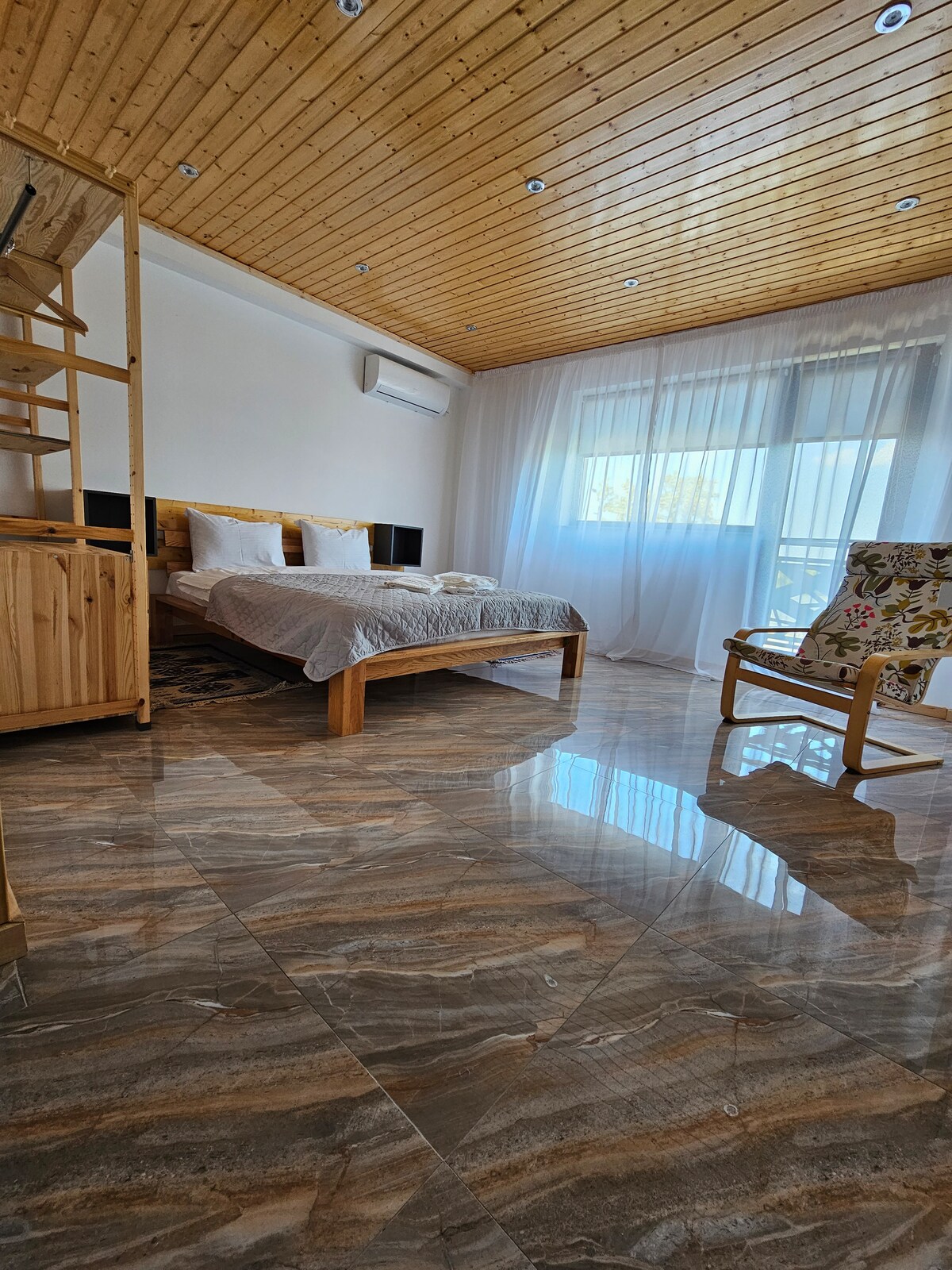 A bright room is observed featuring a spacious wooden bed dressed in light linens. Natural light flows through the sheer curtains covering a large window. Polished marble flooring reflects the room's elements, while a wooden armchair adds additional seating.