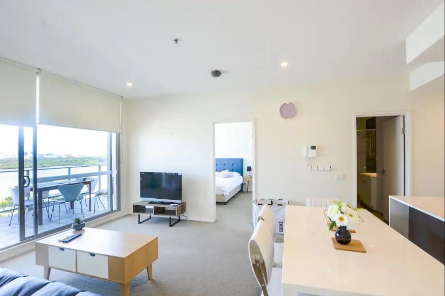 A spacious living area is outlined, featuring a comfortable seating arrangement and a flat-screen television. Natural light filters in through large glass doors that lead to a balcony. A dining table is set for four, while a bedroom is visible in the background.