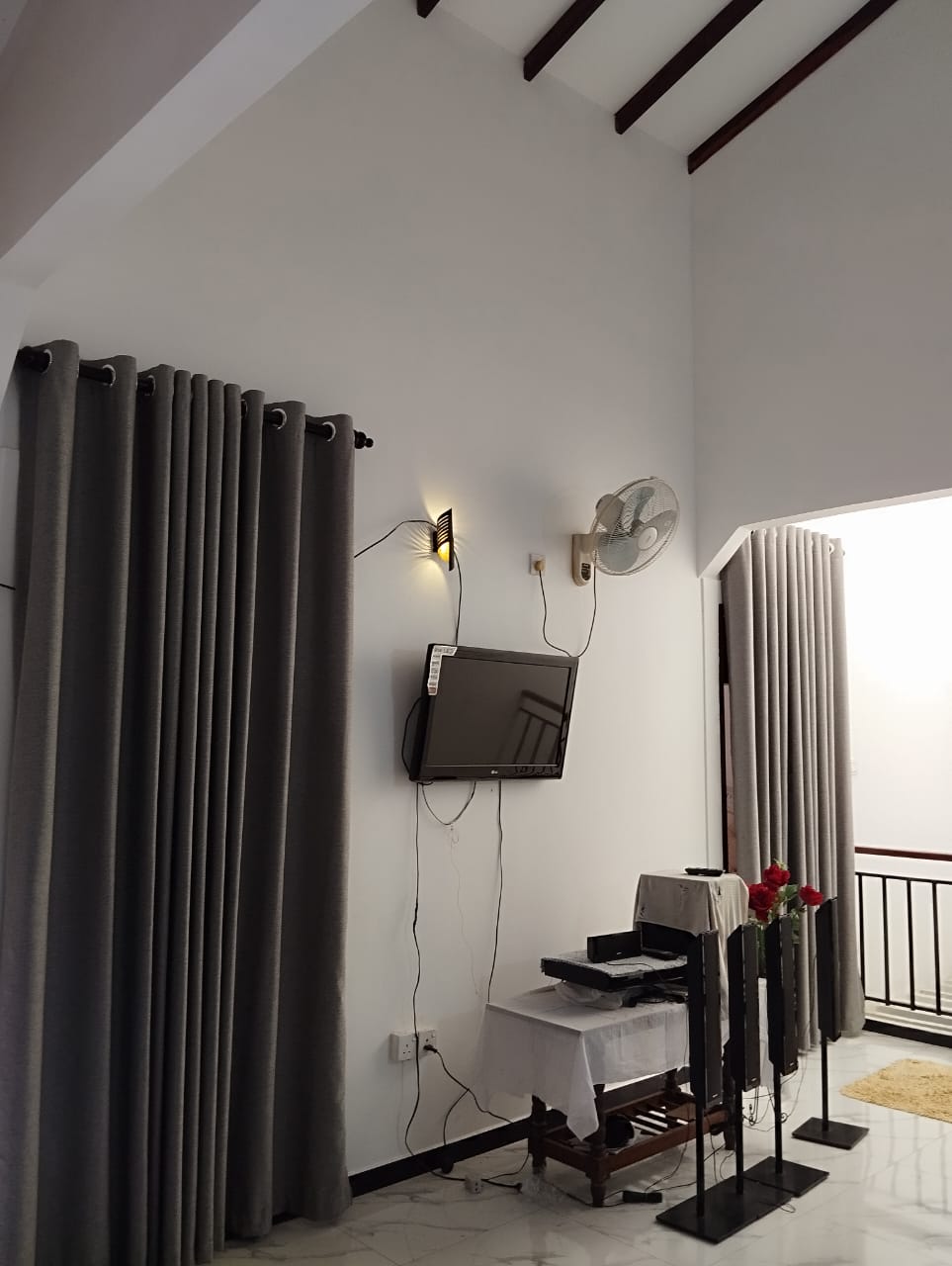 A well-lit corner of the room features a wall-mounted television, with curtains framing the space in soft gray. A small table holds various items, and a decorative fan is mounted on the wall. Light reflects off the glossy tile floor.