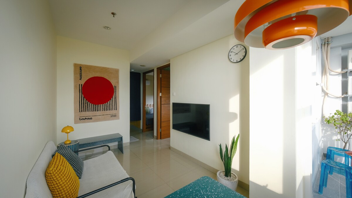A modern living area features a comfortable seating arrangement, complemented by decorative pillows. A vibrant wall art piece with a red circle adds a focal point. Natural light fills the space, while a clock and potted plant enhance the simplicity and style.
