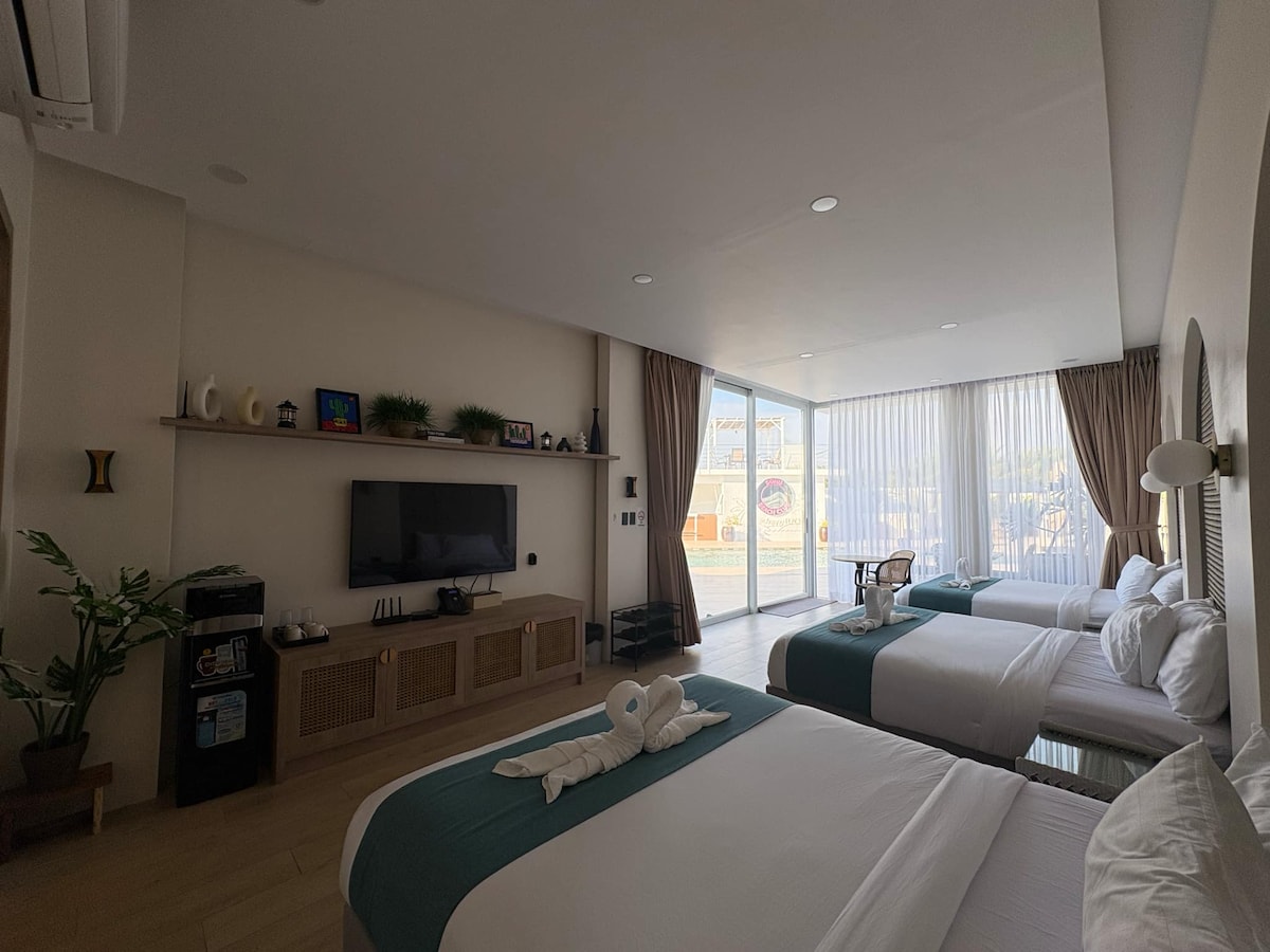 A spacious room is furnished with three beds adorned in crisp white linens and teal accents. A wall-mounted television is positioned across from the beds, complemented by a small refrigerator and a plant. Large glass doors allow natural light and provide access to the outdoor area.