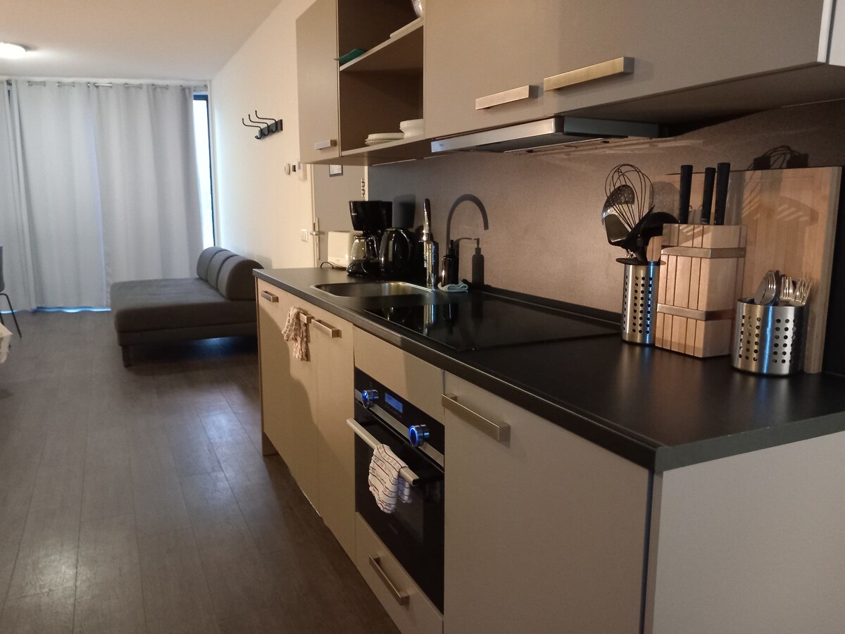 A contemporary kitchen is seen, featuring sleek cabinetry and a dark countertop. Kitchen essentials, including utensils and a coffee maker, are neatly arranged. A cozy sofa is visible in the background, complemented by light coming from the large window covered by soft curtains.