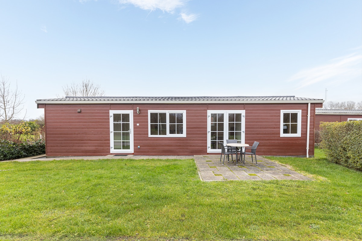 A single-story red wooden cottage is set against a clear sky. The exterior features multiple windows, a pair of double doors, and a small patio area with a table and chairs, surrounded by manicured grass and shrubbery.